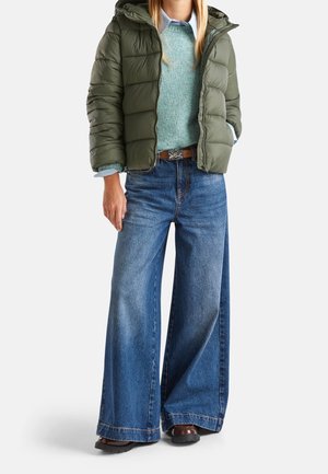 Nainen pukeutuneena oliivinvihreään toppatakkiin, vaaleansiniseen neulepuseroon kauluspaidan päällä, leveälahkeisiin sinisiin farkkuihin ja ruskeisiin kenkiin.