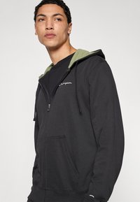 Μαύρο φούτερ με φερμουάρ, με πράσινη εσωτερική κουκούλα, διαθέτει εμπρόσθιες τσέπες, λευκή κεντητή στάμπα λογότυπου και ρυθμιζόμενα κορδόνια.