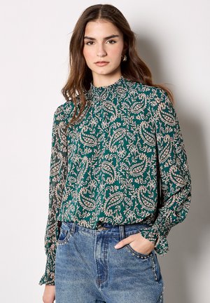Groene blouse met paisleypatroon, een hoge smockhals en lange mouwen, gecombineerd met blauwe denim jeans met decoratieve studs.