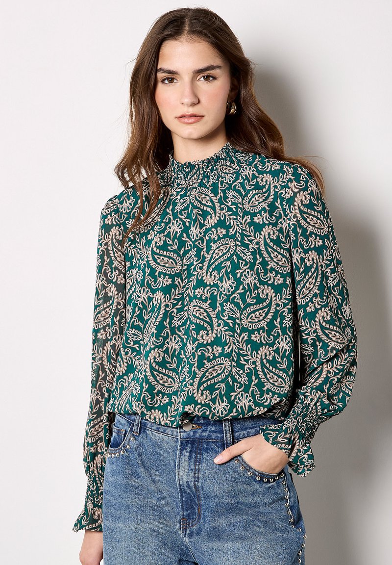 Grüne Bluse mit Paisleymuster, hochgerüschtem Halsausschnitt und langen Ärmeln, kombiniert mit blauen Jeans aus Denim mit dekorativen Nieten.