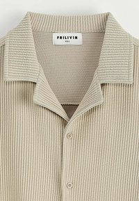 Chemise beige à côtes avec une texture douce, fermeture à boutons et un col. Étiquette indiquant "FRILIVIN, TAILLE S."