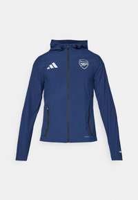 Marineblauwe waterdichte jas met een voorrits, capuchon, Adidas-logo, Arsenal-embleem en zwart ritszakken. Lichtgewicht stof, gladde textuur.
