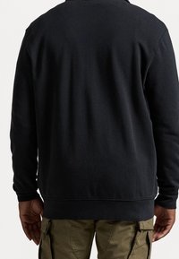 Person som bär en svart långärmad skjorta och olivgröna cargobyxor, sedd bakifrån mot en ljus, enfärgad bakgrund.