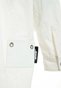 Camisa de algodón blanca con detalles de logo bordado, que presenta una tela texturizada, un bolsillo plano con ojales y etiquetas en negro contrastante.
