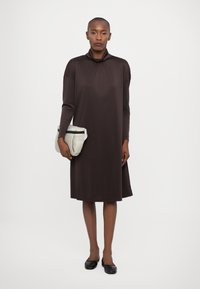 Abito lungo marrone con maniche lunghe e collo a drappeggio, realizzato in tessuto liscio. Indossato con ballerine nere e tiene in mano una piccola borsa color crema.