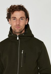 Olivově zelená mikina na zip s vysokým límcem, přední kapsou na zip a nastavitelnými šňůrkami. Tkanina vypadá jemně a má jednoduchý design.