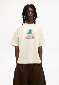 Jasno beżowy, oversize'owy t-shirt z kolorową grafiką palm i różowego samochodu, z krótkimi rękawami i luźnym krojem.