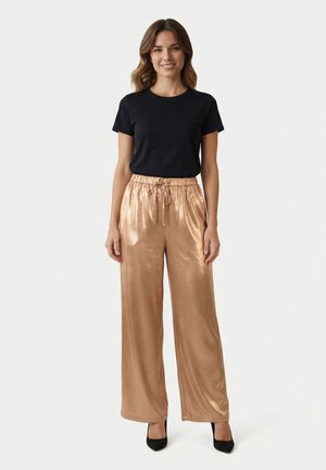 Femme debout, portant un chemisier noir à manches courtes, un pantalon large couleur bronze brillant avec un cordon de serrage, et des chaussures à talons hauts noires, souriante.