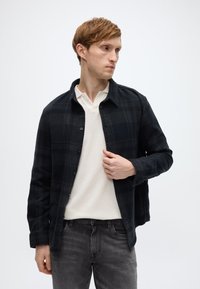 Μαύρο πουκάμισο καρό από φανέλα με σχεδιασμό με κουμπιά, φορεμένο πάνω από ένα πουκάμισο πόλο κρεμ χρώματος. Γκρι τζιν, μινιμαλιστικές λεπτομέρειες.