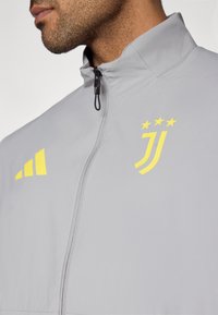 Γκρι μπουφάν με κίτρινες λεπτομέρειες. Διαθέτει ψηλό γιακά, κλείσιμο με φερμουάρ και λογότυπο. Ομαλή υφή και αθλητικός σχεδιασμός.