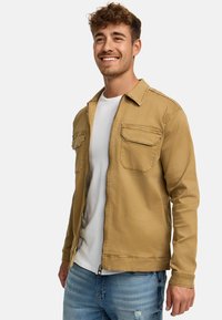 Tan zip-up jakke lavet af bomuld med to brystlommer, krave-design og afslappet pasform. Båret over en hvid t-shirt og lyse jeans.