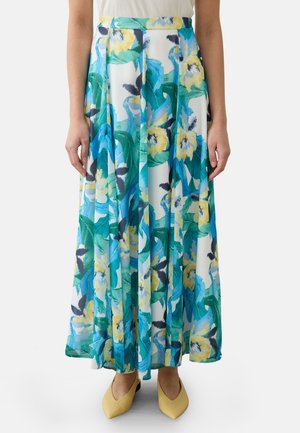 Femme portant une longue jupe fleurie avec des fleurs bleues, vertes et jaunes, assortie à des ballerines pointues jaunes.