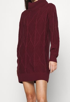 Kobieta w bordowej sukience swetrowej z golfem, wykonanej w technice warkocza, z długimi rękawami oraz prążkowanym wykończeniem na dole i mankietach, widoczna od szyi do połowy ud.