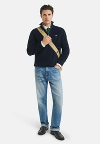 Mann in dunkelblauem Pullover über weißem Hemd mit grün gestreifter Krawatte, hellblauen Jeans, schwarzen Schuhen und beigem Umhängebeutel, der vor einem weißen Hintergrund steht.
