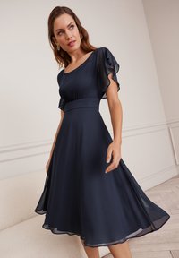 Robe bleu marine avec des manches évasées transparentes, un corsage ajusté et une jupe évasée. Fabriquée en tissu léger au toucher doux.