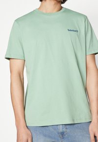 T-shirt en coton vert menthe à manches courtes avec un col rond et un petit logo Timberland bleu sur la poitrine gauche.
