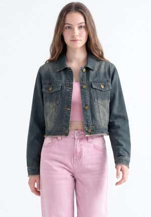 Junge Frau trägt eine ausgebleichte schwarze kurze Jeansjacke über einem rosa gerippten Crop-Top und hoch taillierten rosa Jeans, stehend vor weißem Hintergrund.