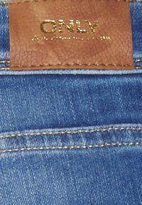 Tecido de ganga em azul claro com costuras escuras, apresentando uma etiqueta de couro castanho embutida com a inscrição "ONLY" em dourado.