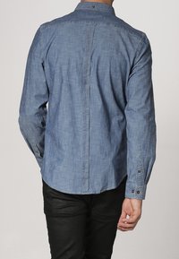 Camisa de mezclilla azul claro con cuello abotonado, mangas largas y costura recta en la parte posterior, con detalles de botones marrones en los puños.