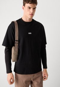 T-shirt nera in cotone con un logo bianco "HUGO" sul petto. Il modello indossa una borsa a tracolla marrone con una tracolla testurizzata e un anello sul dito.