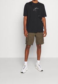 Svart t-shirt med vit text, olivgröna cargoshorts, vita sneakers med grå accenter och svart axelväska. Inga ytterligare detaljer.