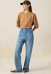 Chemise marron à manches courtes avec des embellissements dorés, jean taille haute bleu clair et une casquette à imprimé animal vert. Chaussures texturées avec un motif en cuir de serpent.