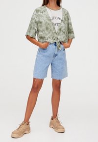 Chemise courte tie-dye verte, T-shirt blanc avec le texte « California », short en denim bleu clair et baskets beiges.