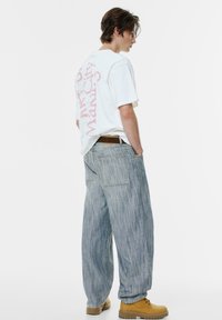 Hvid grafisk T-shirt med pink tekst, løstsiddende blå stribede jeans og brune støvler. Bemærkelsesværdige detaljer inkluderer syningen på baglommen.