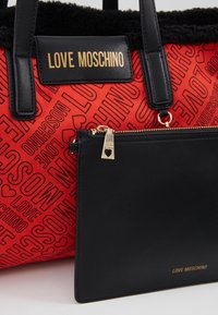 Röd tygväska med svarta läderdetaljer, med texten "LOVE MOSCHINO" i ett upprepande mönster. Inkluderar en avtagbar svart dragkedjefodral.