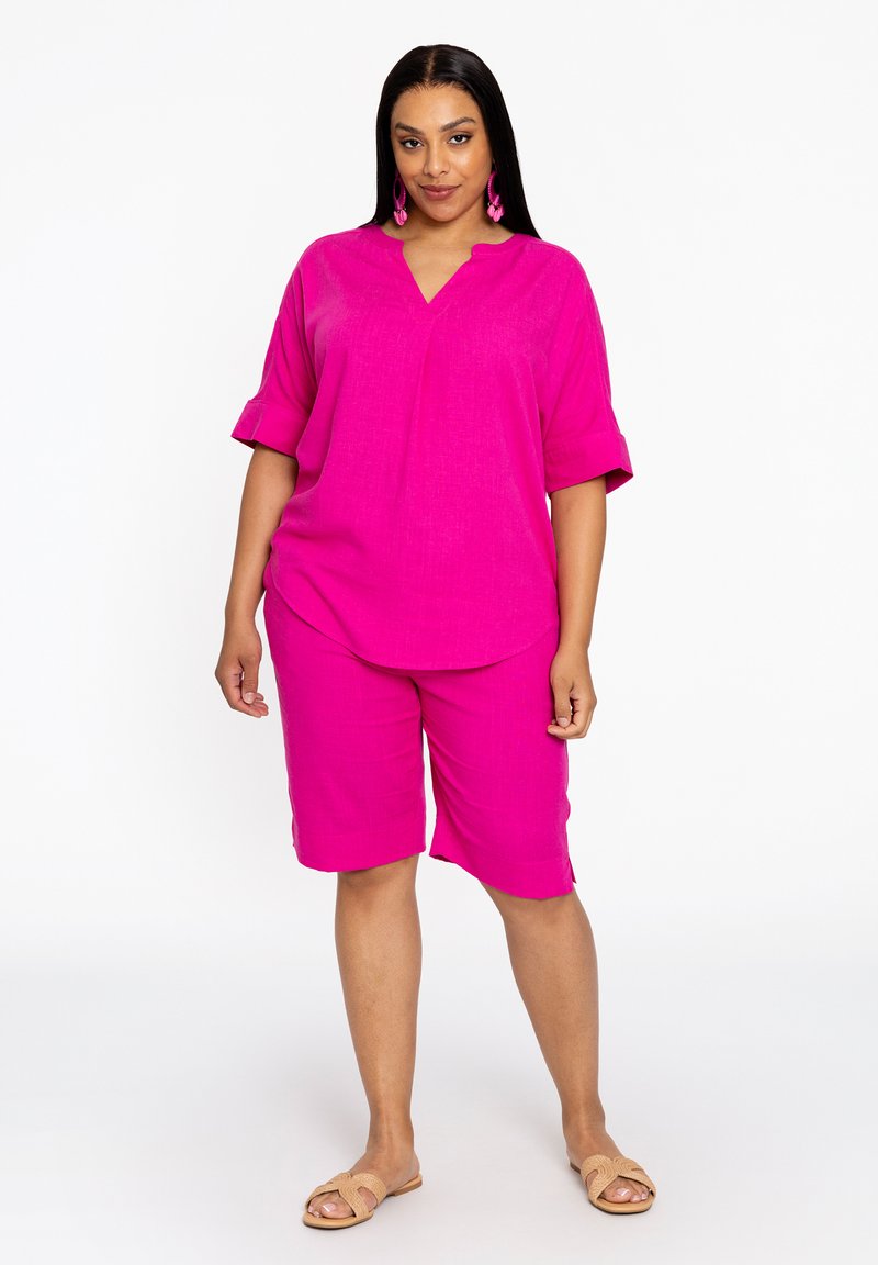 Chemise fuchsia à manches courtes et col en V associée à un short assorti. Tissu léger avec une texture subtile. Portée avec des sandales à glissière beige.