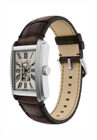 Montre rectangulaire en argent avec un cadran squelette, des chiffres romains noirs, un bracelet en cuir marron et un dessous beige avec des détails de couture.