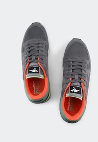 Graue Sneakers mit einem strukturierten Mesh-Obermaterial, orangenem Innenraum, grünen Akzenten an der Ferse und schwarzer Markenkennzeichnung auf der Zunge. Mehrmaterialdesign.