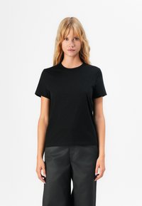 Donna bionda con capelli mossi, che indossa una semplice maglietta nera a maniche corte e pantaloni ampi scuri, in piedi davanti a uno sfondo bianco.