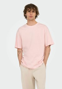 Young man with curly hair wearing a loose light pink T-shirt and beige pants, standing with hands in pockets on a plain background.