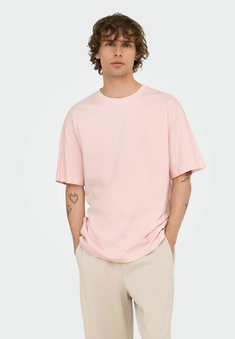 Young man with curly hair wearing a loose light pink T-shirt and beige pants, standing with hands in pockets on a plain background.