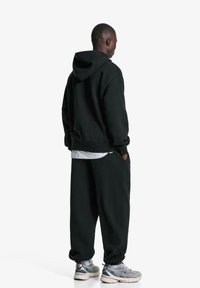 Čierna mikina s kapucňou a zodpovedajúce teplákové nohavice, ktoré majú voľný strih, mäkkú textúru a bočné vrecká, doplnené o šedé športové topánky.
