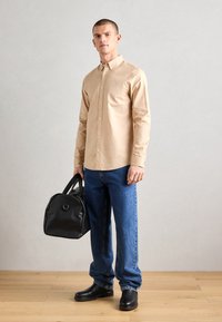 Chemise beige à boutons, jeans bleus, chaussures noires et sac noir. La chemise a un col et un petit détail brodé.