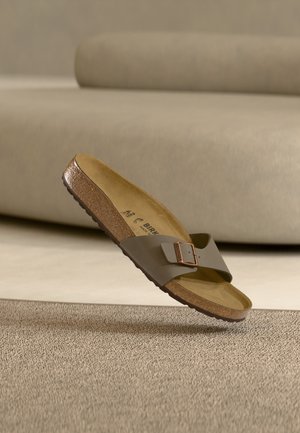 Enkele bruine Birkenstock-sandaal met een gespband die boven een beige tapijt zweeft in een minimalistische kamer met een beige bank.