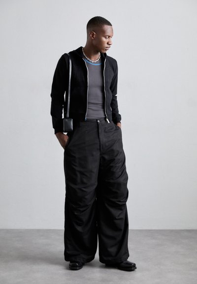 Black zip-up cropped jacket with a hood, gray fitted top, wide black trousers, and black shoes. Small black crossbody bag.