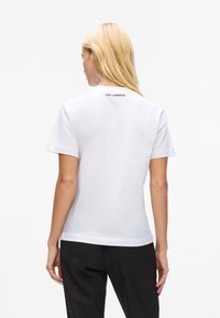 Eine Frau mit langen blonden Haaren steht mit dem Rücken zur Kamera, trägt ein weißes Kurzarmshirt mit dem Text "KARL LAGERFELD" am Hals und schwarze Hosen.