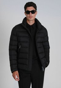 Doudoune noire avec col haut, conception matelassée horizontale, poches latérales zippées et poignets à boutons, portée sur une chemise noire.