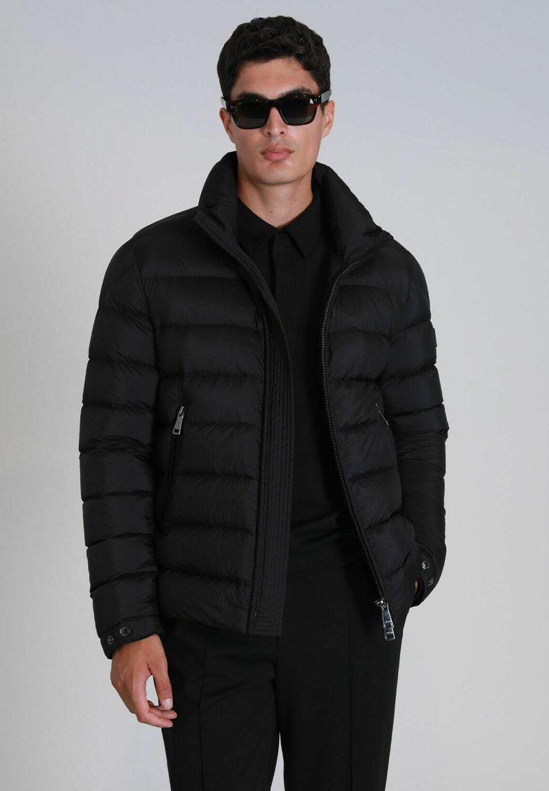 Doudoune noire avec col haut, conception matelassée horizontale, poches latérales zippées et poignets à boutons, portée sur une chemise noire.