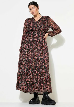 Langes schwarzes Blumenkleid mit rosa und grünen Mustern, langen Ärmeln und einem gestuften Saum. Kombiniert mit klobigen schwarzen Knöchelstiefeln.