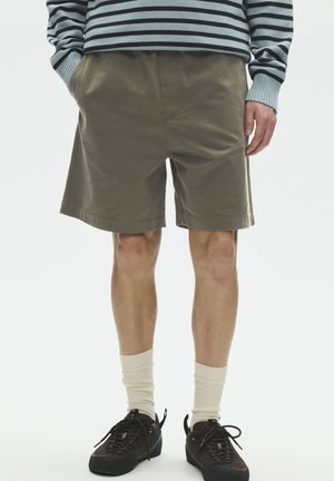 Mand iført olivengrønne shorts, stribet langærmet skjorte, cremefarvede sokker og sorte sneakers, stående med hænderne i lommerne.