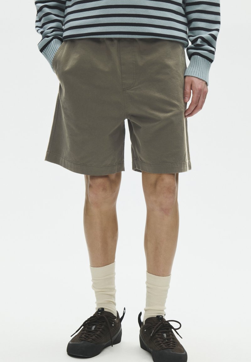 Homme portant un short vert olive, une chemise à manches longues rayée, des chaussettes crème et des baskets noires, debout les mains dans les poches.