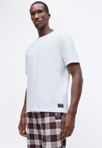 Άνδρας με ένα απλό λευκό κοντομάνικο πουκάμισο και μαρουνί-άσπρα καρό παντελόνια, στέκεται μπροστά σε λευκό φόντο.