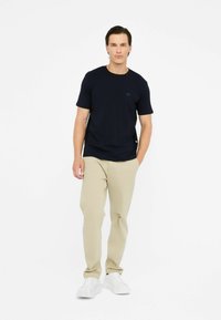 Maglietta a maniche corte blu navy con scollatura rotonda e dettagli logo discreti, abbinata a pantaloni beige chiaro e sneakers bianche.