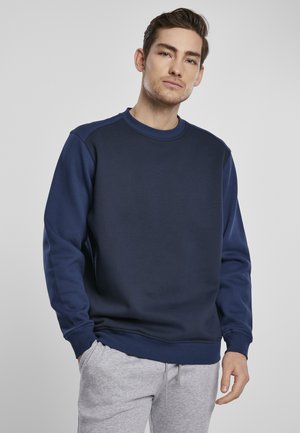 Marineblå sweatshirt med rund hals, struktureret stof og ribbede manchetter. Kombineret med lysegrå sweatpants, der delvist er synlige ved taljen.