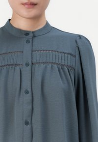 Nærbilde av en person som har på seg en blå-grå bluse med mandarin krage, frontknapper, blonde og plisserte detaljer over brystet.