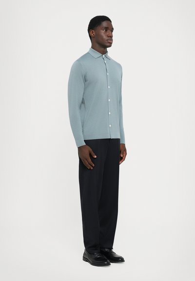 Camisa de botão de manga longa azul claro com colarinho, combinada com calças escuras e sapatos pretos, apresentando uma textura suave e um design ajustado.